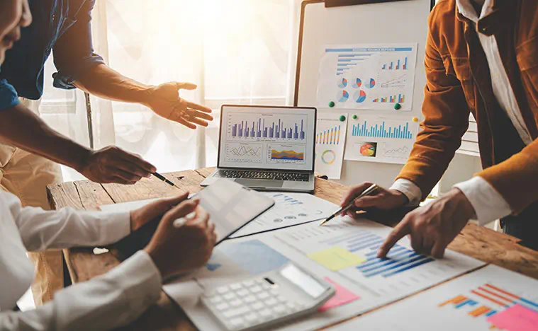 Several business people look over a laptop and a stack of reports.