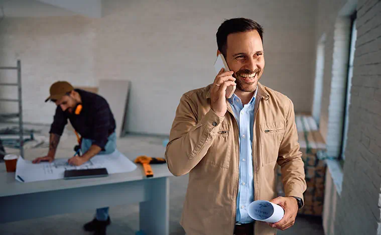 A business man takes a phone call while an architect works in the background.