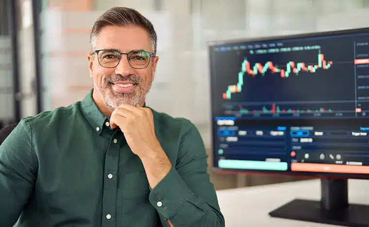 A business man smiles at the camera with his computer monitor showing charts and graphs behind him.