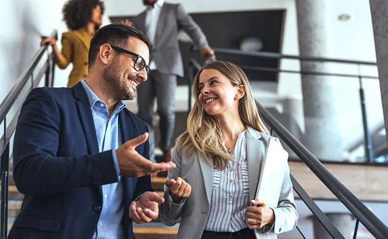 Needs Assessment Two business people talk casually while walking down stairs in a modern office building.