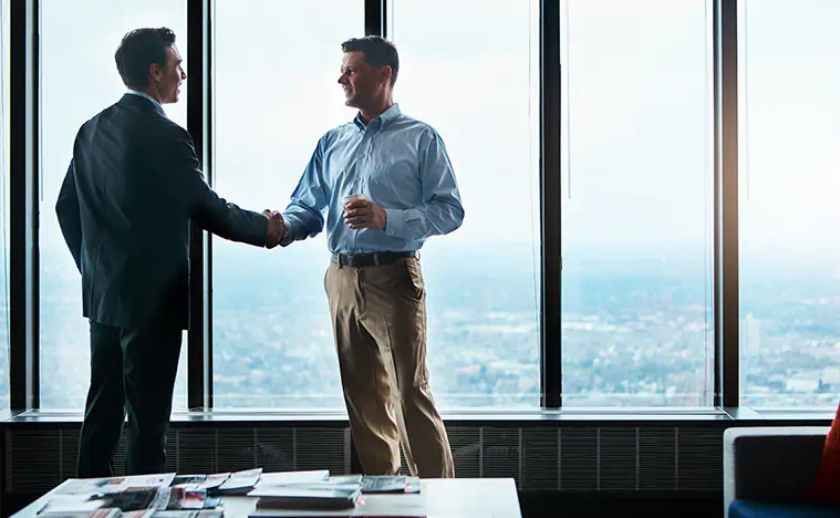 Ongoing Support Two business men shake hands in front of a large glass window of an office.