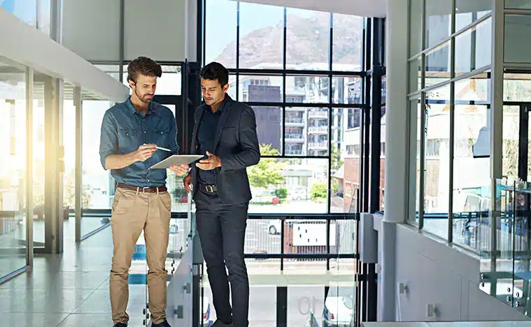 About Us – Communicate Expertly Two business men looking over a tablet in a modern office lobby,